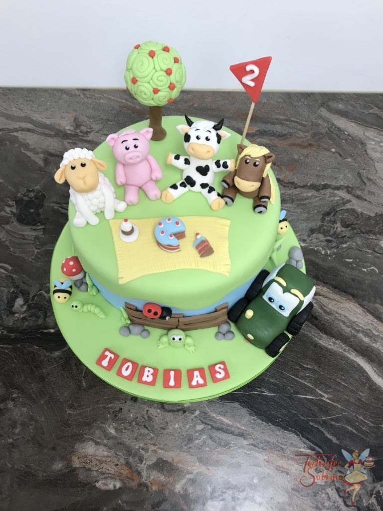 Geburtstagstorte Buben - Bauernhoftiere auf der Wiese, sitzen vergnügt rund um eine Decke mit Leckereien. Ebenfalls auf der Torte ist ein Traktor.
