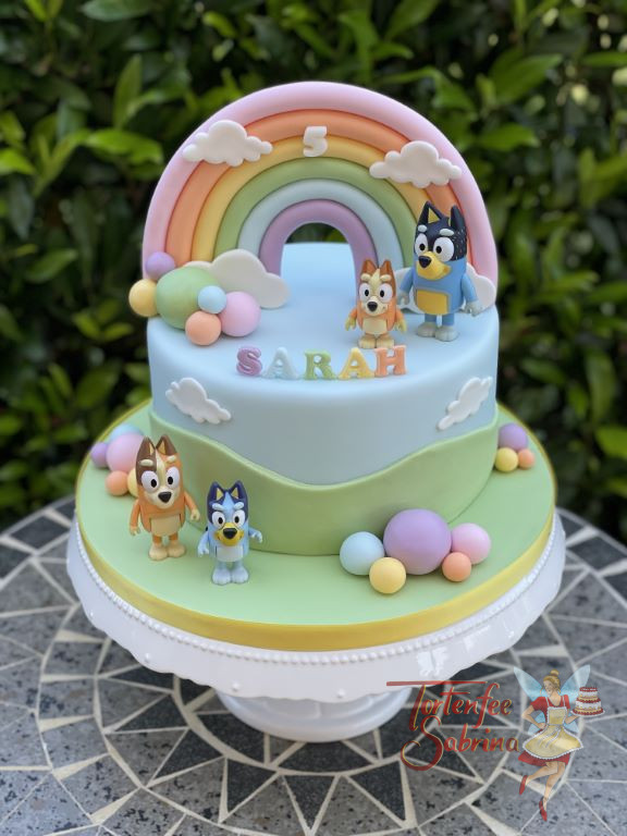 Geburtstagstorte Mädchen - Bluey mit seiner Familie steht auf der Torte und bewundert den bunten Regenbogen.