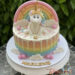 Geburtstagstorte Mädchen - Das lächelnde Einhorn sitz ganz oben auf der Torte über dem Regenbogenmuster und der Wolken.