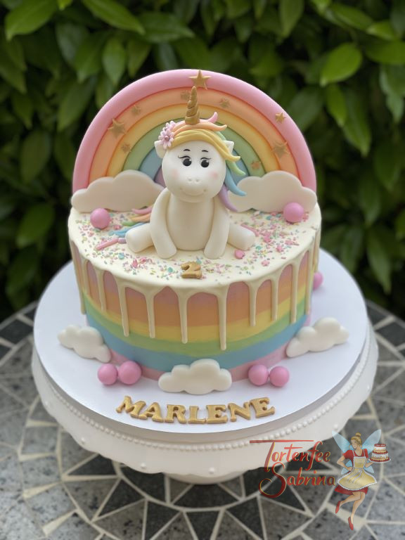 Geburtstagstorte Mädchen - Das lächelnde Einhorn sitz ganz oben auf der Torte über dem Regenbogenmuster und der Wolken.