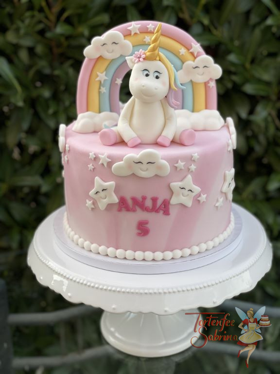 Geburtstagstorte Mädchen - Das sitzende Einhorn ist glücklich und umgeben von einem Regenbogen und Wolken sowie Sternen.