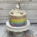 Geburtstagstorte Mädchen - Einhorn auf der Regenbogentorte. Es sitzt unter einer Wimpelkette und zwischen zwei Wolken.