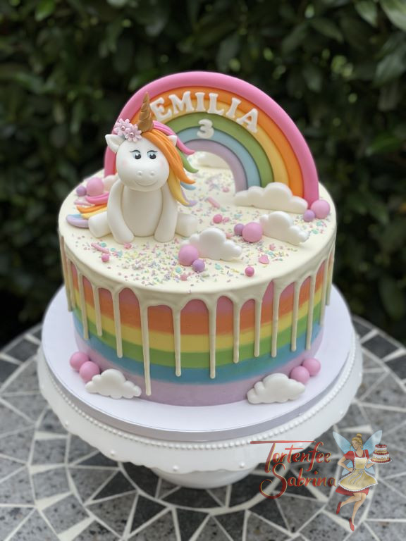 Geburtstagstorte Mädchen - Einhorn auf Wolken und weißem Drip. Die Regenbogenfarben zieren auch die Seite der Torte.