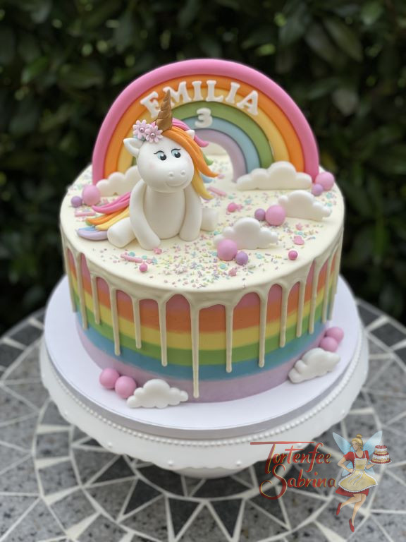 Geburtstagstorte Mädchen - Einhorn auf Wolken und weißem Drip. Die Regenbogenfarben zieren auch die Seite der Torte.