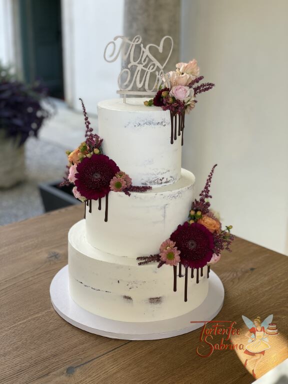 Hochzeitstorte - Dunkelrote Blumen schmücken mit zahlreichen anderen Blumen die Torte, ebenso auf der Torte ist ein Drip aus Schokolade.