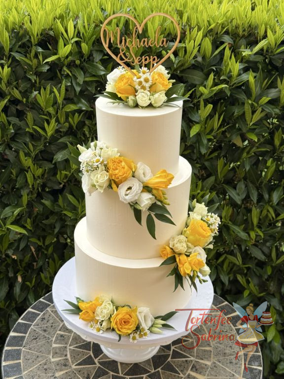 Hochzeitstorte - Herz aus Holz mit den Namen des Brautpaares verziert ganz oben die weiße dreisttöckige Torte neben den Blumenschmuck.