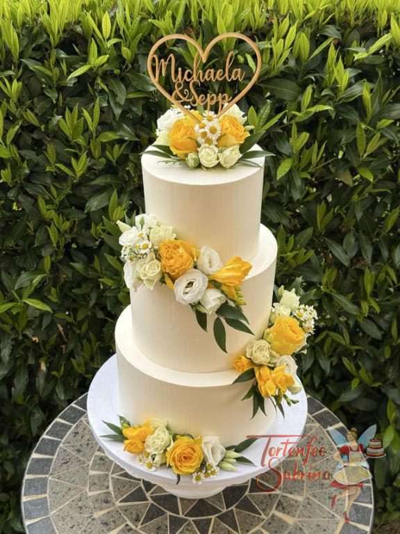 Hochzeitstorte - Herz aus Holz mit den Namen des Brautpaares verziert ganz oben die weiße dreisttöckige Torte neben den Blumenschmuck.