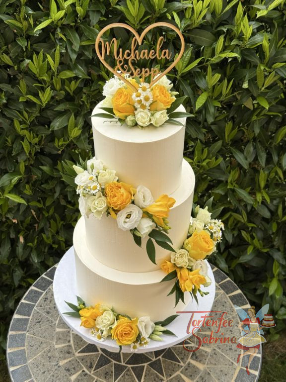 Hochzeitstorte - Herz aus Holz mit den Namen des Brautpaares verziert ganz oben die weiße dreisttöckige Torte neben den Blumenschmuck.