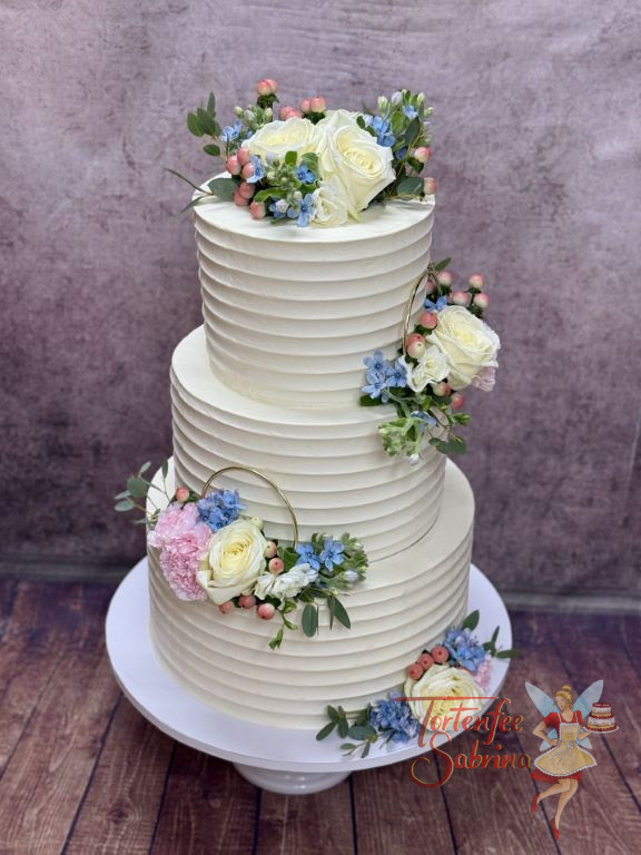 Hochzeitstorte - Weiße und blaue Blüten verzieren neben den goldenen Ringen und den exacten Rillen die dreistöckige Torte.