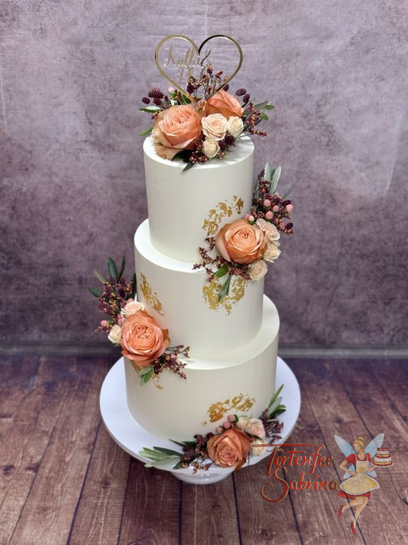 Hochzeitstorte - Herz mit Schrift in goldener Farbe ziert ganz oben die weiße dreistöckige Torten neben Blumen und Blattgold.